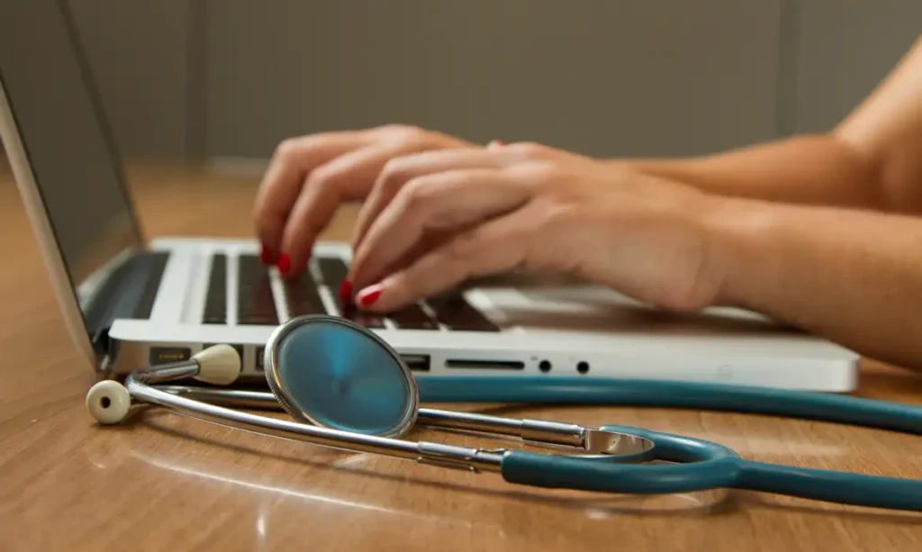 Médica digita no computador, tendo seu estetoscópio ao lado. Foto: National Cancer Institute/Unsplash