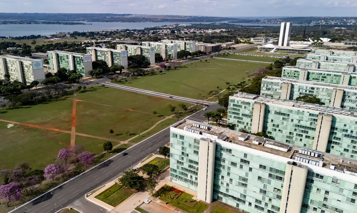 22/06/2026 - Brasília - A floração dos ipês-roxos transforma a paisagem de Brasília em um espetáculo de cores que dura apenas algumas semanas, mas deixa marcas na memória durante todo o ano. Considerada uma das árvores mais simbólicas do Cerrado brasileiro, o ipê anuncia a chegada da estação seca justamente quando perde suas folhas e exibe flores em tons vibrantes que contrastam com o céu azul da Esplanada dos Ministérios. Em meio às linhas retas, ao concreto aparente e aos tons cinza característicos da arquitetura modernista idealizada por Oscar Niemeyer, os ipês surgem como protagonistas da paisagem urbana. A explosão de flores roxas quebra a monotonia das formas e colore avenidas, parques e áreas residenciais, criando uma das cenas mais marcantes da capital federal. Breve e intensa, a floração dos ipês lembra que alguns dos mais belos espetáculos da natureza são também os mais passageiros. Foto: Rafa Neddermeyer/Agência Brasil